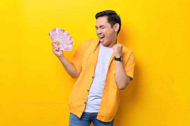 Opgewonden jonge knappe Aziatische man die een hoop geld vasthoudt en succes viert geïsoleerd op gele achtergrond