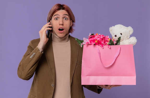 Opgewonden jonge knappe man die aan de telefoon praat en een roze cadeauzakje met bloemen en teddybeer vasthoudt