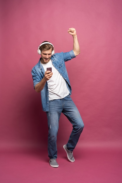 Opgewonden jonge man met een spijkerbroek, geïsoleerd over een roze achtergrond, luisterend naar muziek met oortelefoons en mobiele telefoon