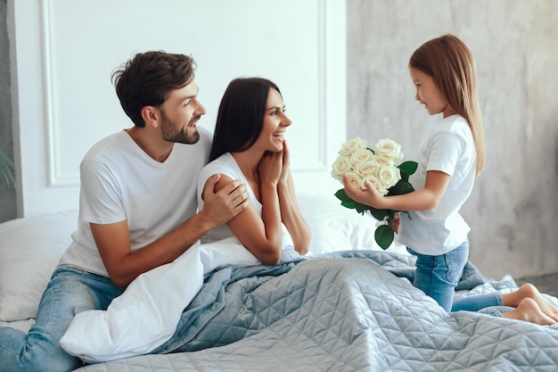 Opgewonden jonge ouders kijken hoe een klein meisje een bos witte rozen naar hen brengt terwijl ze op een bed zitten
