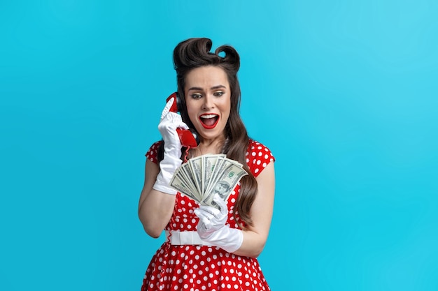 Opgewonden jonge pinup-vrouw in rode polka dot-jurk met geld dat op de vaste telefoon spreekt op blauw