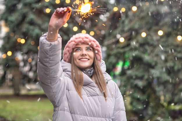 Foto opgewonden jonge vrouw draagt roze gebreide muts en jas die plezier heeft met sterretjes op straat bij de kerstboom tijdens de sneeuwval