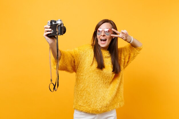 Opgewonden jonge vrouw in hart brillen doen selfie geschoten op retro vintage fotocamera met overwinning teken geïsoleerd op gele achtergrond. Mensen oprechte emoties, levensstijl. Reclame gebied.