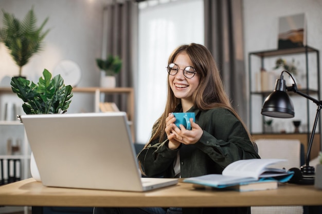 Opgewonden jonge vrouw met beker met warme drank en laptop voor videogesprek