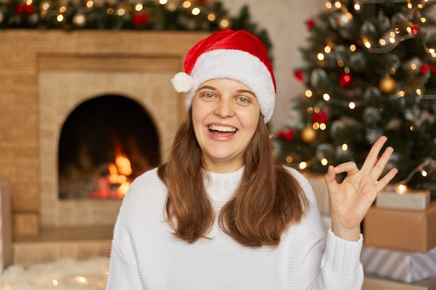 Opgewonden jonge vrouw met kerstman hoed en trui gebaren ok teken, met opwinding, haar tanden laten zien, poseren binnen met open haard en kerstboom.