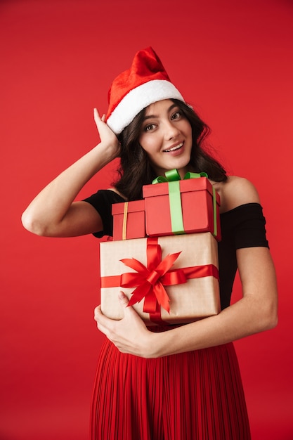 Opgewonden jonge vrouw met kerstmuts staande geïsoleerd over rood, met stapel geschenkdozen