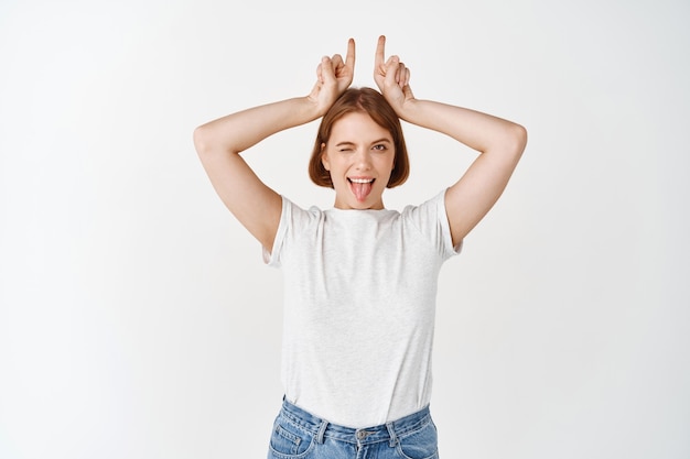 Opgewonden jonge vrouw met tong en hoorns teken, blij knipogen, staande in casual t-shirt en jeans op witte muur