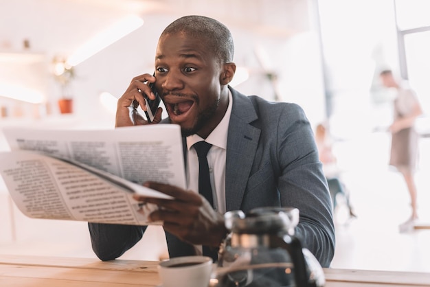 Opgewonden knappe Afro-Amerikaanse zakenman met smartphone krant lezen tijdens de koffiepauze