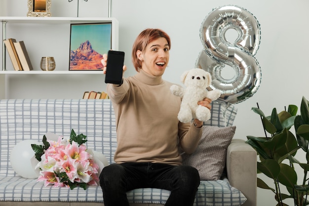 Opgewonden knappe man op gelukkige vrouwendag met teddybeer met telefoon zittend op de bank in de woonkamer