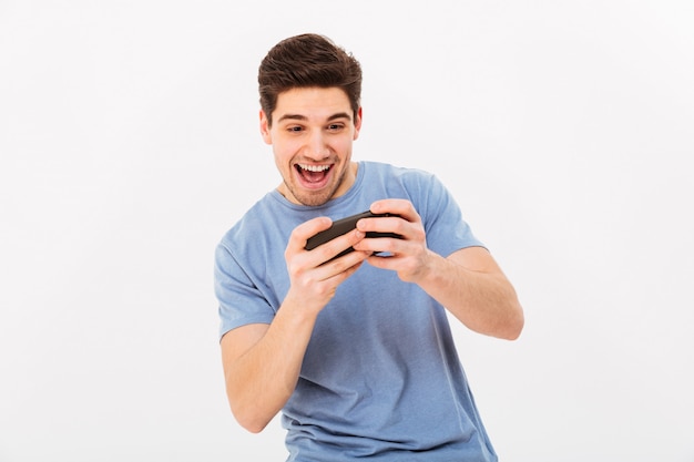 Opgewonden man in casual t-shirt spelen van online games op smartphone, geïsoleerd over witte muur