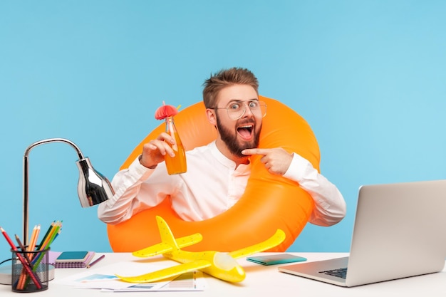 Opgewonden man kantoormedewerker zittend op de werkplek met rubberen reddingsboei en speelgoedvliegtuig wijzende vinger naar fles met limonade, ontspannend. Indoor studio-opname geïsoleerd op blauwe achtergrond