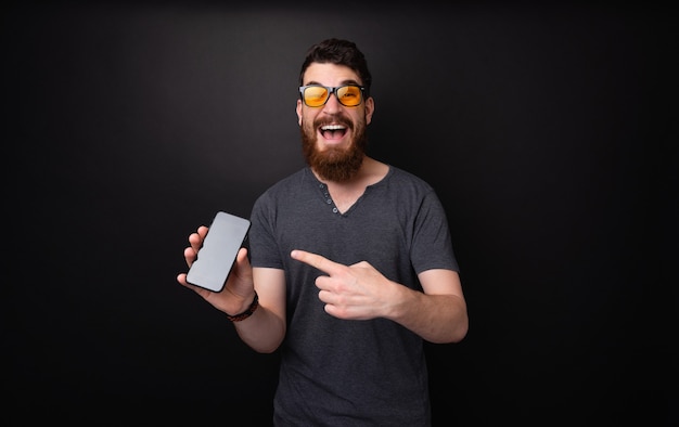 Opgewonden man met baard en zonnebril wijzend op mobiele telefoon over donkere geïsoleerde achtergrond