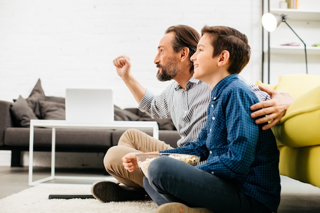 Opgewonden man zit met zijn gelukkige zoon en een vuist opsteken tijdens het kijken naar een geweldige sportwedstrijd op tv thuis