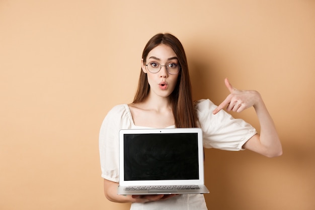 Opgewonden meisje in glazen presentatie maken op computer, hand wijzend op laptop scherm en zeggen wow, staande op beige achtergrond.