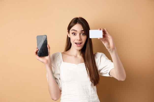 Opgewonden meisje met plastic creditcard van bank en leeg gsm-scherm, demonstreren shopping app, staande op beige.