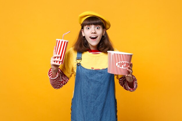 Opgewonden meisje tiener in franse baret, denim zomerjurk met plastic beker cola of frisdrank, emmer popcorn geïsoleerd op gele achtergrond. mensen oprechte emoties, lifestyle concept. bespotten kopie ruimte.