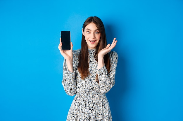 Opgewonden meisje toont leeg smartphonescherm en hijgend gefascineerd, demonstreren shopping-app, staande op blauw.