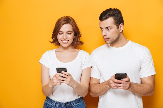 Opgewonden mooi stel met witte t-shirts die geïsoleerd over een gele muur staan en mobiele telefoons vasthouden