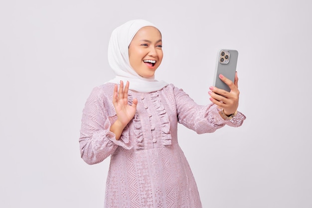 Opgewonden mooie jonge Aziatische moslimvrouw die hijab en paarse jurk draagt met behulp van een smartphone die een videogesprek voert of selfie neemt geïsoleerd op een witte studioachtergrond