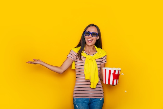 Opgewonden mooie moderne jonge brunette vrouw in vrijetijdskleding en 3D-bioscoopbril en grote mand popcorn poseert tijdens het kijken naar een film in de bioscoop