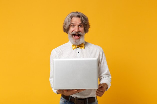 Opgewonden oudere grijsharige snor bebaarde man in wit overhemd vlinderdas geïsoleerd op geel oranje achtergrond, studio portret. Mensen levensstijl concept. Bespotten kopie ruimte. Bedrijf laptop pc-computer.