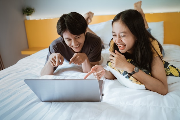 Opgewonden paar lachen op bed met behulp van de laptop thuis in de slaapkamer samen