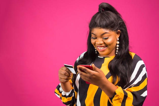 Opgewonden PlusSize Afrikaanse vrouw met behulp van telefoon en creditcard poseren over roze Studio achtergrond mobiel winkelen concept