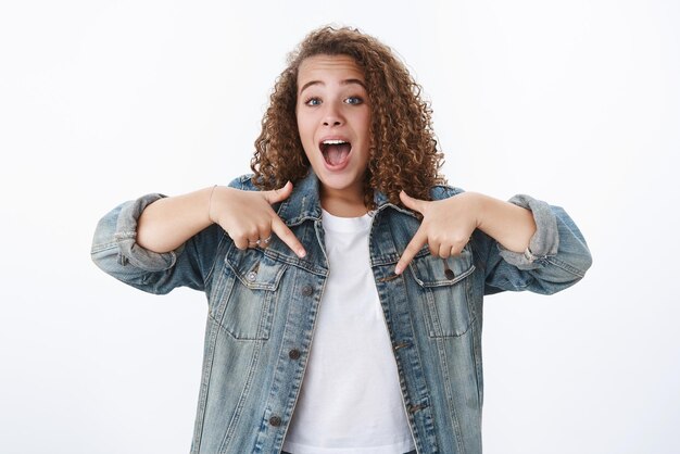 Opgewonden positieve charismatische gekrulde Europese vrouw blauwe ogen schreeuwen opgewonden uitleggen waar vind plaats wijzend naar beneden wijsvingers uitchecken cool product witte achtergrond dragen van denim