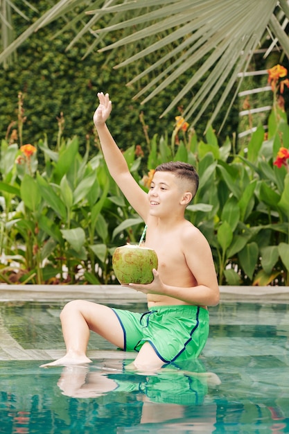 Opgewonden preteenjongen die in zwembad met kokoscocktail zit en met hand zwaait om de aandacht van vriend te vestigen
