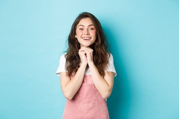 Opgewonden romantisch jong meisje, hand in hand in de buurt van de borst en kijken met anticipatie en tederheid, glimlachen, nadenken over iets moois, blauwe achtergrond