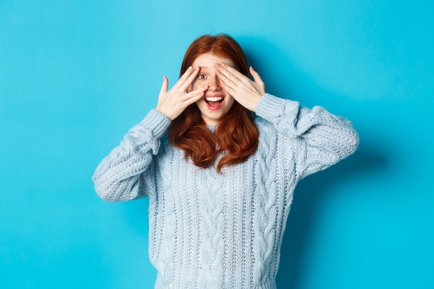 Opgewonden roodharige tienermeisje opent ogen om vakantie verrassing te zien, cadeautjes ontvangen, camera verbaasd kijken, staande over blauwe achtergrond.