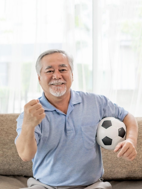 Opgewonden senior man oude man voelt zich gelukkig met voetbal voetbal bereid je voor op cheer team favoriet op de bank van de bank thuis levensstijl aziatische senior goede gezondheid concept