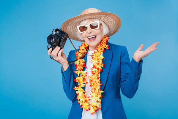 Opgewonden senior vrouw reiziger met retro fotocamera geïsoleerd op blauwe achtergrond