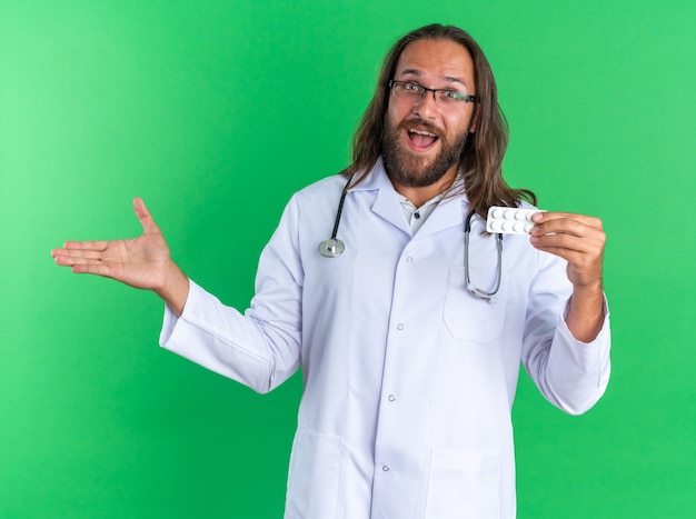 Opgewonden volwassen mannelijke arts met een medisch gewaad en een stethoscoop met een bril die naar de camera kijkt met een pak tabletten en lege hand geïsoleerd op een groene muur