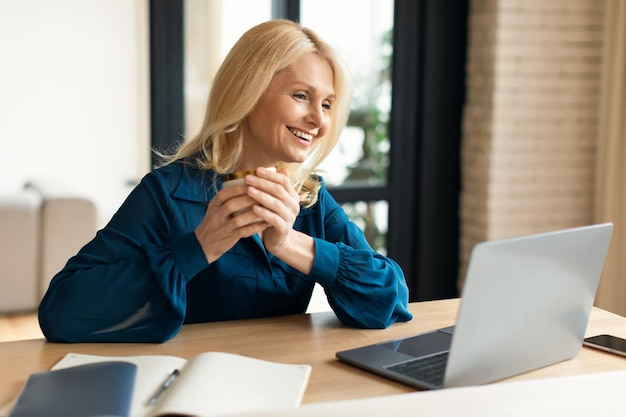 Opgewonden volwassen zakenvrouw die online werkt op een laptop en koffie drinkt terwijl ze op de werkplek zit