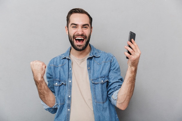 Opgewonden vrolijke man met shirt geïsoleerd over grijze muur, met behulp van mobiele telefoon, vieren