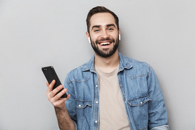 Opgewonden vrolijke man met shirt geïsoleerd over grijze muur, met oortelefoons, met behulp van mobiele telefoon