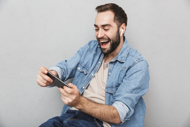 Opgewonden vrolijke man met shirt geïsoleerd over grijze muur, met oortelefoons, met behulp van mobiele telefoon