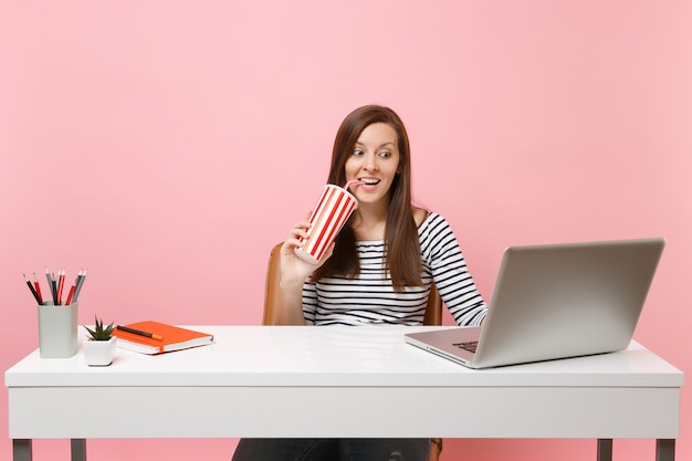 Opgewonden vrouw die drinkt uit een plastic beker met cola-frisdrank zit te werken aan een project op kantoor aan een wit bureau met een moderne pc-laptop