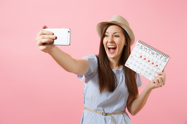 Opgewonden vrouw in blauwe jurk doet selfie op mobiele telefoon, met periodenkalender voor het controleren van menstruatiedagen geïsoleerd op roze achtergrond. medisch, gezondheidszorg, gynaecologisch concept. ruimte kopiëren.