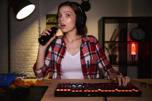 Opgewonden vrouw met hoofdtelefoon spelen van online games op computer, snacks eten