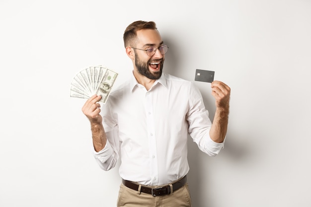 Opgewonden zakenman geld houden en kijken naar creditcard, staande op witte achtergrond.