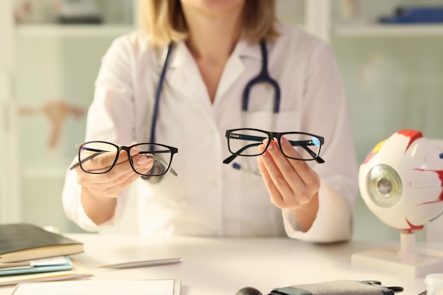 Ophthalmologist holds different eyeglasses in her hands while sitting at desk in medical clinic