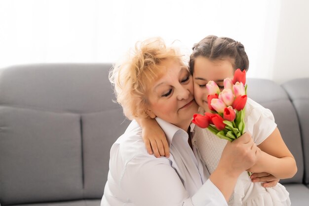 Oprechte schattige kleine kleindochter sterk knuffelen en zoenen in de wang haar 60s grootmoeder gaf haar mooie lentebloemen feliciteert met verjaardag, Internationale Vrouwendag, close-up concept afbeelding.