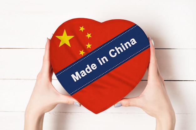 Opschrift Made in China, de vlag van China. Vrouwelijke handen met een hartvormige doos. Witte houten tafel op een muur. Plaats voor tekst