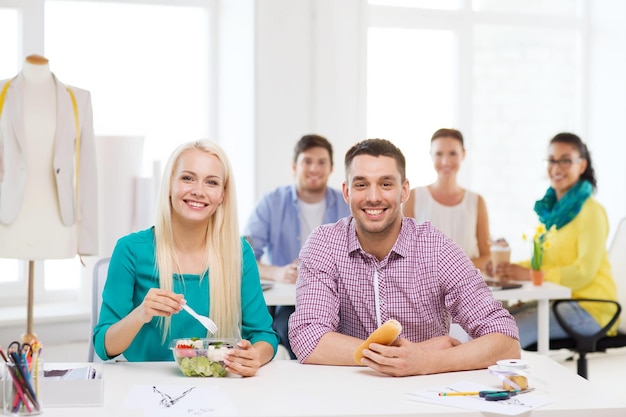 opstarten, onderwijs, mode, eten en kantoorconcept - glimlachende modeontwerpers die lunchen op kantoor