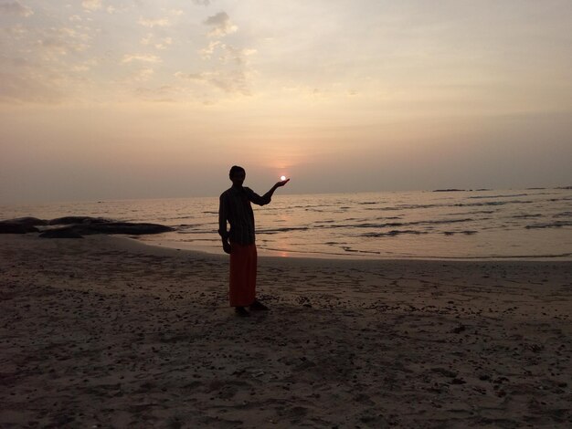 Photo optical illusion of silhouette man holding sun while standing at beach against sky