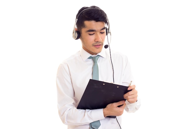 Optimisitic young man with black hair in white shirt with blue tie and headphones holding a folder on white background in studio