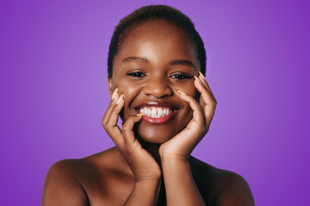 Optimistic african american woman smiling while touching perfect skin of cheek isolated over purple background advertisement for skin cream antiwrinkle cream baby face