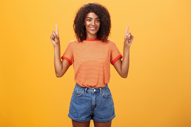 Optimistic ambitious stylish dark-skinned female in striped cool t-shirt and shorts raising hands pointing upwards.